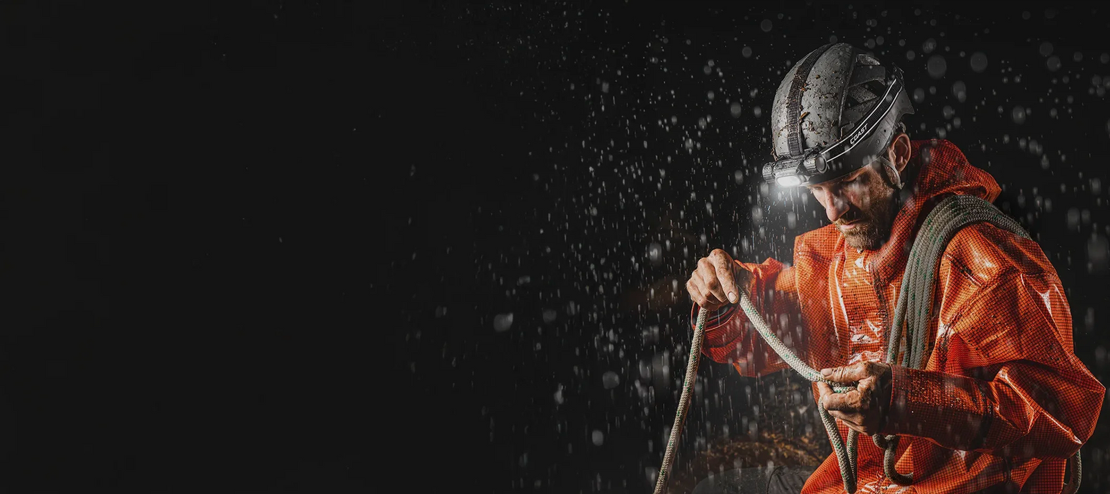 Tehokas Coast otsalamppu kypärään kiinnitettynä ja mies työskentelee narun kanssa vesisateessa
